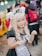 A person dressed in a maid costume with cat ears, featuring long blonde hair and a playful expression. They are surrounded by a crowd of people, some wearing backpacks.
