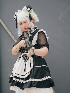 A person dressed in a black and white stylized costume with lace and ribbon details. They have white hair styled with ornate accessories and are holding a firearm. The person's expression is focused and serious, with visible tattoos on their arms.