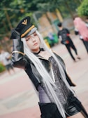A person dressed in a detailed cosplay costume with a long silver wig and a stylish black outfit including a military-style hat with gold accents. The background shows blurred figures of other individuals, possibly in a park or outdoor setting.