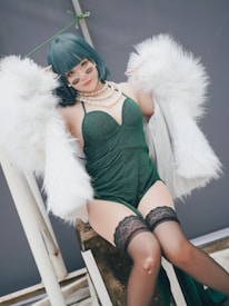 A person wearing a stylish emerald green dress and stockings, accessorized with round sunglasses and a layered pearl necklace. The outfit is complemented by a luxurious white faux fur coat draped over the shoulders. The individual has dark green bobbed hair and is sitting on a stone-like surface, posing confidently against a muted background.