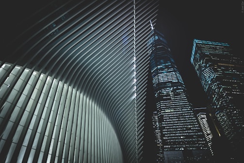 A futuristic architectural scene featuring the interior of a building with sleek, curved ribs and two adjacent skyscrapers with numerous lit windows against a night sky.