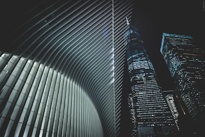 A futuristic architectural scene featuring the interior of a building with sleek, curved ribs and two adjacent skyscrapers with numerous lit windows against a night sky.