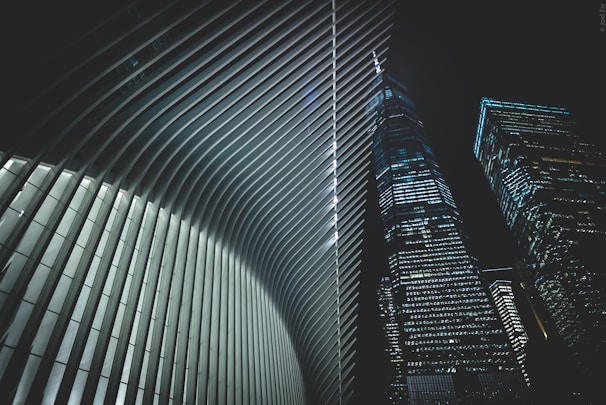A futuristic architectural scene featuring the interior of a building with sleek, curved ribs and two adjacent skyscrapers with numerous lit windows against a night sky.