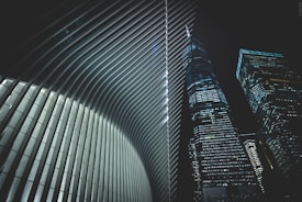 A futuristic architectural scene featuring the interior of a building with sleek, curved ribs and two adjacent skyscrapers with numerous lit windows against a night sky.