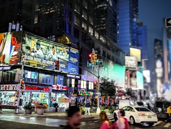 A bustling city street at night featuring bright billboards and LED screens with advertisements. The scene includes various retail stores and a red traffic light at an intersection. Pedestrians and vehicles, including a white car, populate the street.