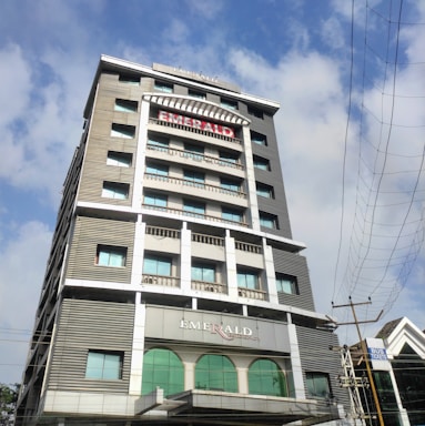 A tall multi-story building named Emerald Residency is shown with a modern architectural design, featuring large windows and a prominent sign at the top. The building is surrounded by a clear blue sky with some clouds and nearby power lines.