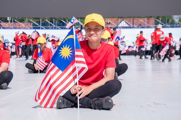 A group of individuals, most of them dressed in red shirts and yellow caps, are seated on the ground holding the Malaysian flag. In the foreground, a person is sitting cross-legged and smiling while holding the flag. More people can be seen in the background holding flags and standing in a semicircle, with a large structure or stage in the distance.