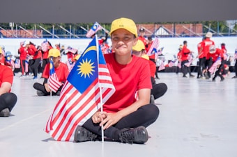 A group of individuals, most of them dressed in red shirts and yellow caps, are seated on the ground holding the Malaysian flag. In the foreground, a person is sitting cross-legged and smiling while holding the flag. More people can be seen in the background holding flags and standing in a semicircle, with a large structure or stage in the distance.