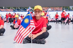 A group of individuals, most of them dressed in red shirts and yellow caps, are seated on the ground holding the Malaysian flag. In the foreground, a person is sitting cross-legged and smiling while holding the flag. More people can be seen in the background holding flags and standing in a semicircle, with a large structure or stage in the distance.
