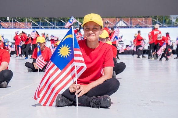 A group of individuals, most of them dressed in red shirts and yellow caps, are seated on the ground holding the Malaysian flag. In the foreground, a person is sitting cross-legged and smiling while holding the flag. More people can be seen in the background holding flags and standing in a semicircle, with a large structure or stage in the distance.