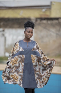 A person stands outdoors wearing a flowing garment with a patterned design featuring earth tones. The garment has a prominent central stripe of solid color, and the background includes a blurred, neutral-colored wall.