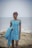 A person stands on a rocky shoreline with a calm sea in the background. They are wearing an off-the-shoulder blue dress with a floral pattern and holding a matching piece of fabric in one hand.