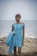 A person stands on a rocky shoreline with a calm sea in the background. They are wearing an off-the-shoulder blue dress with a floral pattern and holding a matching piece of fabric in one hand.