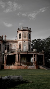 An old, weathered mansion with a tall, cylindrical tower and a wrap-around porch supported by columns. The building's facade shows signs of decay, with peeling paint and crumbling bricks. Set against a cloudy sky, it is surrounded by a grassy lawn and trees.