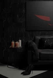 A dark and moody living room setting featuring a black sofa with a decorative pillow, a small round side table with three lit candles of different sizes, and a framed abstract artwork on the wall. The floor is partially visible with high-heeled shoes placed neatly. The color palette is predominantly dark, with touches of red in the artwork.