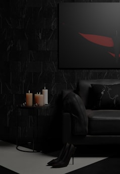 A dark and moody living room setting featuring a black sofa with a decorative pillow, a small round side table with three lit candles of different sizes, and a framed abstract artwork on the wall. The floor is partially visible with high-heeled shoes placed neatly. The color palette is predominantly dark, with touches of red in the artwork.