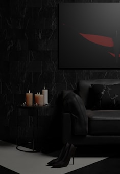 A dark and moody living room setting featuring a black sofa with a decorative pillow, a small round side table with three lit candles of different sizes, and a framed abstract artwork on the wall. The floor is partially visible with high-heeled shoes placed neatly. The color palette is predominantly dark, with touches of red in the artwork.