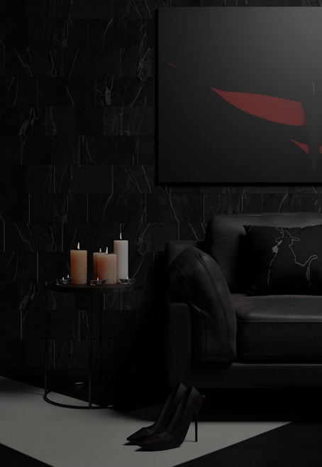 A dark and moody living room setting featuring a black sofa with a decorative pillow, a small round side table with three lit candles of different sizes, and a framed abstract artwork on the wall. The floor is partially visible with high-heeled shoes placed neatly. The color palette is predominantly dark, with touches of red in the artwork.