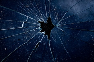 A close-up view of a shattered glass surface with multiple angular cracks radiating out from a central point. The glass appears slightly dirty with dark, shadowy reflections. The color palette is predominantly dark blue and black, adding a moody and dramatic effect.