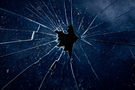 A close-up view of a shattered glass surface with multiple angular cracks radiating out from a central point. The glass appears slightly dirty with dark, shadowy reflections. The color palette is predominantly dark blue and black, adding a moody and dramatic effect.