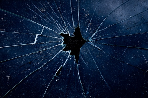 A close-up view of a shattered glass surface with multiple angular cracks radiating out from a central point. The glass appears slightly dirty with dark, shadowy reflections. The color palette is predominantly dark blue and black, adding a moody and dramatic effect.