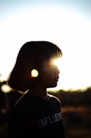 A silhouette of a person with short hair captured against a bright, setting sun. The light creates a halo effect around her head, and there is a notable lens flare in the image. The background is blurred and the sunlit horizon provides a warm ambiance.