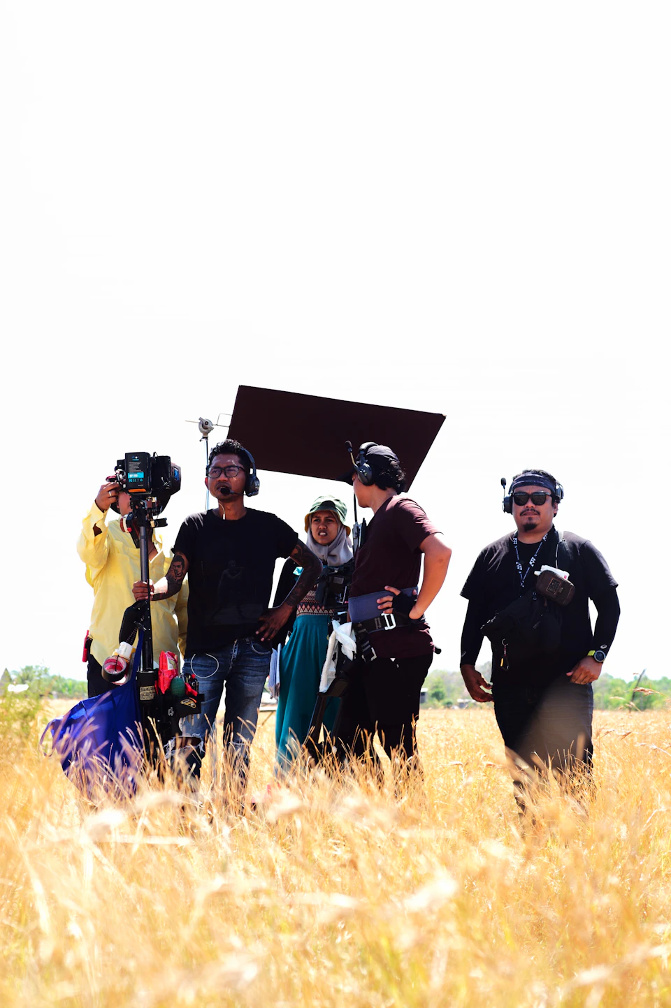 The image shows a group of five people standing in a field, with their backs to the camera and facing away from it. The person on the far left is holding a camera, while another individual stands next to them wearing headphones. In the center, there's someone holding a large black object that appears to be a piece of equipment or a prop for filming. On the right side, two more individuals are present, one with a backpack and the other carrying a microphone. The field they're standing in is filled with tall grasses, creating an outdoor setting for their gathering.
