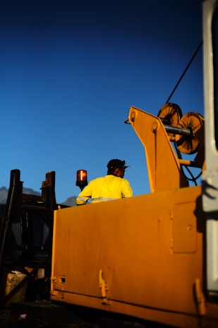 A worker wearing a yellow jacket and cap is standing on a large industrial machine, possibly a crane, under a clear blue sky. The machine features bright yellow components and a prominent lifting mechanism.
