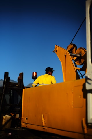 A worker wearing a yellow jacket and cap is standing on a large industrial machine, possibly a crane, under a clear blue sky. The machine features bright yellow components and a prominent lifting mechanism.