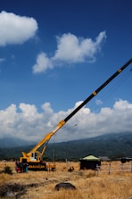 A yellow crane is positioned in an open field with a backdrop of mountains and blue sky. The crane's boom is extended at an angle, and a few people are gathered near its base. A small green tent is visible to the right, along with some fencing and dry grass covering the ground. White clouds are scattered across the sky.