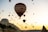 Numerous hot air balloons fill the sky during dawn or dusk, creating a picturesque scene. The largest balloon with text on it dominates the foreground, while others are scattered across the sky. Rugged terrain and natural rock formations lie below, partially covered in vegetation.