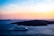 A cruise ship sails on a vast blue sea, capturing the serene beauty of the scene. In the background, dark islands are silhouetted against a stunning gradient sky, transitioning from pale blue to warm orange and pink hues. The calm water reflects the soft evening light, enhancing the tranquil atmosphere.