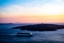 A cruise ship sails on a vast blue sea, capturing the serene beauty of the scene. In the background, dark islands are silhouetted against a stunning gradient sky, transitioning from pale blue to warm orange and pink hues. The calm water reflects the soft evening light, enhancing the tranquil atmosphere.