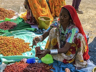 A woman wearing colorful traditional clothing and silver bangles is seated on the ground, sorting through small fruits. She has a pile of red and yellow berries in front of her on a green fabric and is placing some of them into a clear container. Other women, also in vibrant dresses, are partially seen in the background. A yellow bucket and a blue cup are nearby.