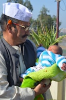 A man wearing a white cap and glasses is gently holding a newborn baby swaddled in a green blanket and dressed in a blue and white knitted outfit. A small flower is tucked behind the man's ear, and there is a mark on his forehead. They are outdoors, with green plants and a clear blue sky in the background.