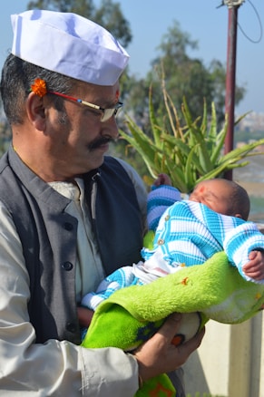 A man wearing a white cap and glasses is gently holding a newborn baby swaddled in a green blanket and dressed in a blue and white knitted outfit. A small flower is tucked behind the man's ear, and there is a mark on his forehead. They are outdoors, with green plants and a clear blue sky in the background.
