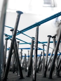An artistic perspective focusing on numerous metal nails embedded vertically into a surface. Blue threads are intricately wrapped around the nails, creating an abstract pattern. The background is blurred, highlighting the nails and threads.