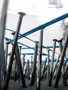 An artistic perspective focusing on numerous metal nails embedded vertically into a surface. Blue threads are intricately wrapped around the nails, creating an abstract pattern. The background is blurred, highlighting the nails and threads.