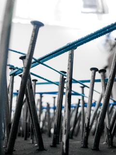 An artistic perspective focusing on numerous metal nails embedded vertically into a surface. Blue threads are intricately wrapped around the nails, creating an abstract pattern. The background is blurred, highlighting the nails and threads.