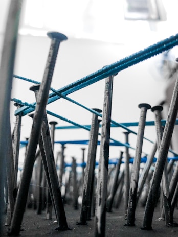 An artistic perspective focusing on numerous metal nails embedded vertically into a surface. Blue threads are intricately wrapped around the nails, creating an abstract pattern. The background is blurred, highlighting the nails and threads.