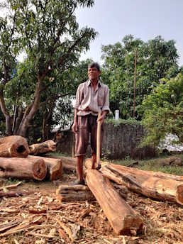 A man stands on a pile of cut logs holding an axe, surrounded by large trees and greenery. The scene appears to be outdoors in a natural setting, with pieces of wood and bark scattered on the ground.