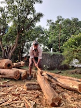 A person chopping wood outdoors surrounded by cut logs and wooden shavings. Lush green trees and a rustic stone wall are in the background, indicating a natural, rural setting.
