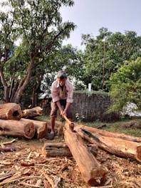A person chopping wood outdoors surrounded by cut logs and wooden shavings. Lush green trees and a rustic stone wall are in the background, indicating a natural, rural setting.