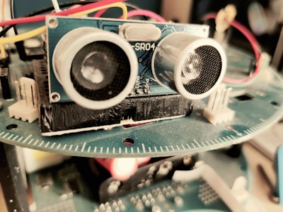 A close-up view of an electronic device featuring a blue circuit board with components such as sensors and wires. The focus is primarily on an ultrasonic sensor with two circular transducers. Red, yellow, and white wires are connected, and a blurred background suggests it is part of a larger apparatus.
