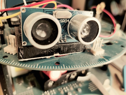 A close-up view of an electronic device featuring a blue circuit board with components such as sensors and wires. The focus is primarily on an ultrasonic sensor with two circular transducers. Red, yellow, and white wires are connected, and a blurred background suggests it is part of a larger apparatus.