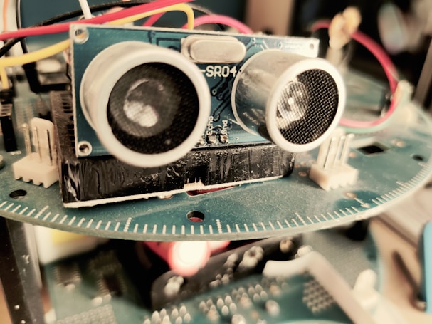 A close-up view of an electronic device featuring a blue circuit board with components such as sensors and wires. The focus is primarily on an ultrasonic sensor with two circular transducers. Red, yellow, and white wires are connected, and a blurred background suggests it is part of a larger apparatus.