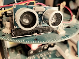 A close-up view of an electronic device featuring a blue circuit board with components such as sensors and wires. The focus is primarily on an ultrasonic sensor with two circular transducers. Red, yellow, and white wires are connected, and a blurred background suggests it is part of a larger apparatus.