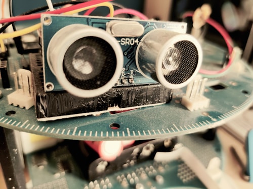 A close-up view of an electronic device featuring a blue circuit board with components such as sensors and wires. The focus is primarily on an ultrasonic sensor with two circular transducers. Red, yellow, and white wires are connected, and a blurred background suggests it is part of a larger apparatus.