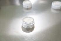 A small open jar of cream stands on a reflective silver surface. The label with the brand name 'PLANTADEA' is visible. The focus is on the product, while other similar jars appear blurred in the background.