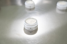 A small open jar of cream stands on a reflective silver surface. The label with the brand name 'PLANTADEA' is visible. The focus is on the product, while other similar jars appear blurred in the background.